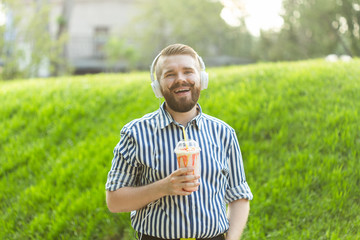 Side view of a stylish young man with a beard holding a milkshake and admiring the city views walking in the park on a warm summer day. The concept of rest and relaxation.