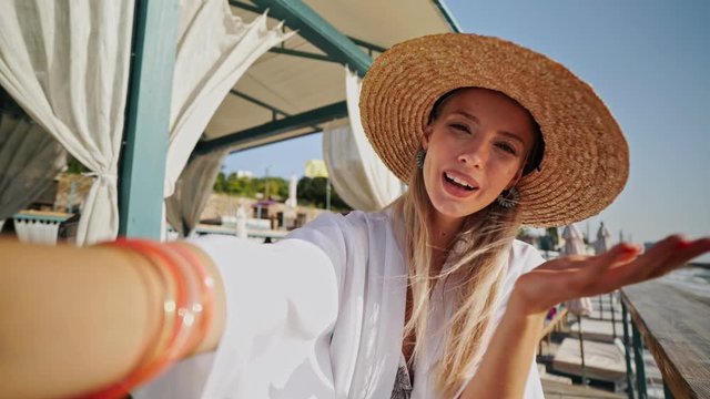 Gorgeous blonde woman 20s in summer straw hat have a rest on the beach take a selfie by camera