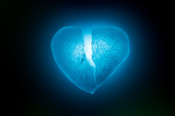 Ice frozen broken in half heart symbol of blue color close-up glowing in the dark in blue cold light. Texture of ice with bubbles. Symbol of a broken heart. Allegory of unhappy love.