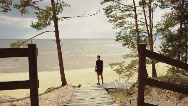 Young Man is Standing on a Wooden Rural Platform on the Edge of a Pine Forest and Looking at a Beautiful Blue Sea Landscape