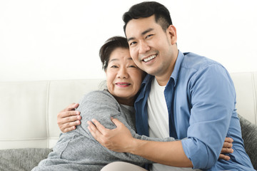 Asian mother hugging her son and sitting on sofa at home, lifestyle concept.