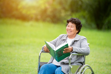 Elderly Asian women, elderly, sit on wheelchairs and read books outdoors in the park.