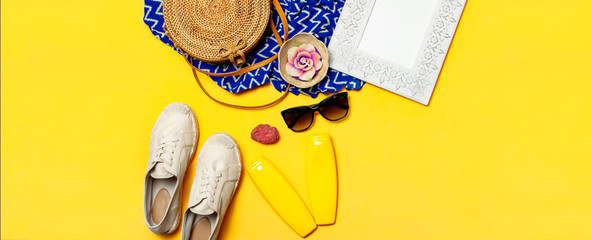 Fototapeta na wymiar Female summer fashion background. Clothes and accessories on yellow background. Blue dress, round rattan bag, espadrilles shoes, sunglasses, sunscreen, wooden picture frame. Flat lay top view