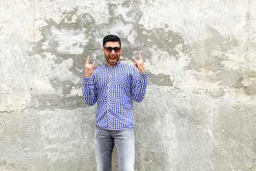 Obraz na płótnie Canvas Portrait of surprised fuuny handsome bearded young man in checkered blue shirt and sunglasses standing against concrete gray wall in rock and roll horns gesture, looking and screaming at camera.