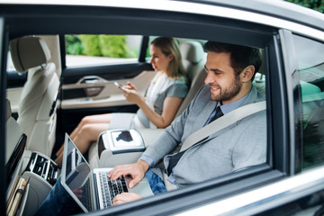 Business couple with laptop sitting on back seats in car, working.