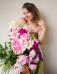 young attractive girl in dress with bouquet of peonies is standing with one hand near hair, looking straight and smiling happy on white wall background, romantic concept, free space