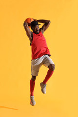 Fototapeta na wymiar Full length portrait of black man in red shirt, black cap and grey shorts playing basketball. Studio, yellow background