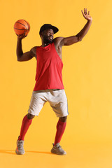 Full length portrait of black man in red shirt, black cap and grey shorts playing basketball. Studio, yellow background