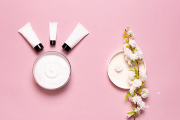 Beauty Spa concept. Opened plastic container with cream, cosmetic bottle containers, spring White flowers on pink background Flat lay top view. Herbal dermatology cosmetic hygienic cream organic