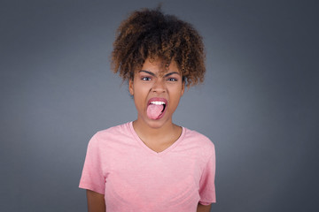 Beautiful African American woman with happy and funny face smiling and showing tongue. Wearing casual clothes and standing against gray studio background.