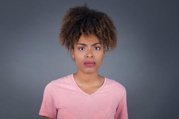 Portrait of outraged young African American woman, and long hair wearing pink t-shirt frowning her eyebrows being displeased with something. Scowling pretty female isolated over grey.ï¿½