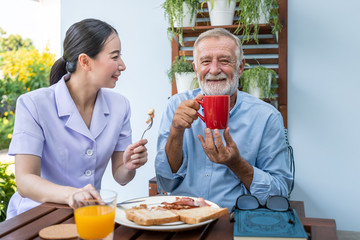 Nurse assist elderly senior man to eat breakfast and drink coffee with mug in hand at nursing home