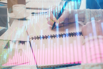 Forex chart hologram on hand taking notes background. Concept of analysis. Double exposure