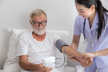 Happy nurse checking and measuring elderly man blood pressure with device in bedroom at nursing home