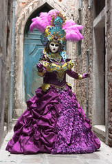 Fototapeta na wymiar Mysterious mask with bright purple dress, adorned with embroidery, gems and feathers on the hat, posing in a typical Venice street during the traditional carnival.