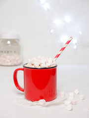 red mug with hot chocolate and a lot of melted little marshmallows. In the background there is a jar withlittle marshmallows . Christmas lights in the background.