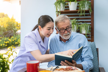 Fototapeta na wymiar Senior elderly man reading book with nurse during breakfast in garden at nursing home