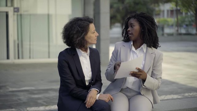 Serious businesswomen sitting on street and discussing contract. Confident business partners communicating while sitting on bench. Business communication concept