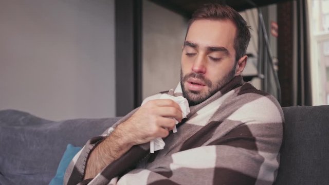 Upset sick bearded man covering in blanket while sitting on sofa at home