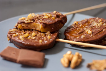 Slices of apples in chocolate, caramel glaze and walnuts on skewers on a dark background
