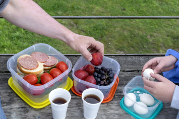 On a wooden table in a box are tomatoes and sandwiches with sausage. The container has fruits such as plums and grapes. Next to the food are cups of tea. The man picked up the fruit.