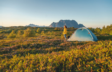 Reizigersmeisje in een geel jasje staat bij zonsondergang naast een tent in Noorwegen