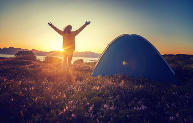 Reisendes Mädchen in einer gelben Jacke steht bei Sonnenuntergang neben einem Zelt in Norwegen