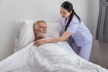 Obraz premium Happy nurse cover elderly man with blanket in bedroom at nursing home