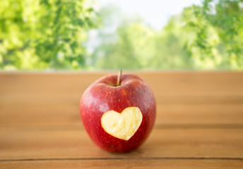 fruits, food and health concept - ripe red apple with carved heart shape on wooden table over green natural background