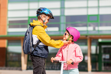 safety, childhood and care concept - happy school boy with backpack fastening girl's helmet for scooter riding
