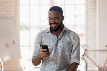 Happy black businessman holding smartphone using apps standing in office
