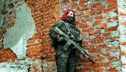Red-hair airsoft woman in uniform with machine-gun, stand beside brick wall.