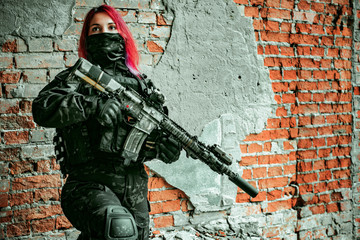 Airsoft soldier red-hair woman in uniform with machine gun inside broken building beside brick wall