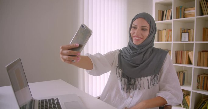 Closeup portrait of young cheerful muslim businesswoman in hijab taking selfies on phone posing sitting in front of laptop in office indoors