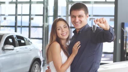 Handsome caucasian man holding car keys in hand posing with his gorgeous girlfriend near new luxury car at the dealership.