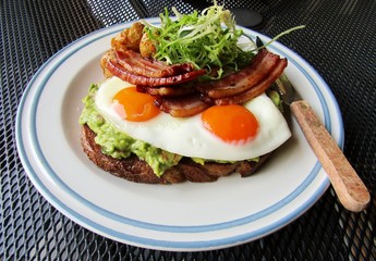 Mashed avocado on whole grain bread toast with sunny side egg topped with bacon, roasted baby potatoes and curly endive lettuce