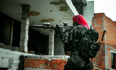 Airsoft red-hair woman in uniform with machine gun standing on knee. Soldier aims at the sight on ruine.