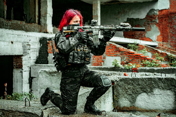 Fototapeta premium Airsoft red-hair woman in uniform with machine gun standing on knee. Soldier aims at the sight on ruine. Side view