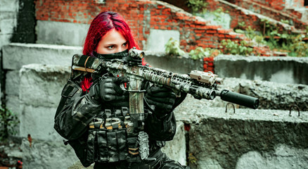 Fototapeta premium Airsoft red-hair woman in uniform with machine gun. Soldier aims at the sight on broken building