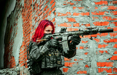 Airsoft red-hair woman in uniform with machine gun beside brick wall. Soldier aims at the sight on the ruins. Close up
