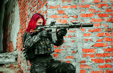 Fototapeta premium Airsoft red-hair woman in uniform with machine gun beside brick wall. Soldier aims at the sight on the ruins. Side view