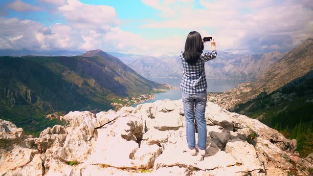 woman traveller standing on the rock take photo view of the mountainous area with blue water houses in the valley surrounded trees