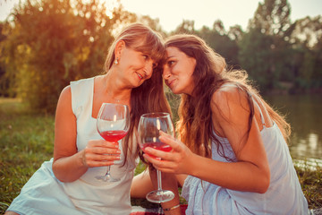 Mother drinking wine with her adult daughter by summer river at sunset. Family having picnic. Mother's day