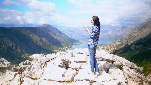 woman traveller standing on the rock scrolls feed social media or sharing photo view of the mountainous area with blue water houses in the valley surrounded trees