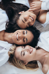 top view of three smiling multiethnic women in white shirts lying with closed eyes