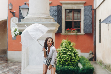 Happy young girl walking around town under umbrella. It's summer rain. Beautiful brunette young woman wearing shirt and shorts.