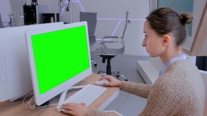 Woman looking at white monitor of desktop computer with blank green display in modern futuristic office interior. Mock up, copyspace, workspace, chroma key, template, green screen, technology concept