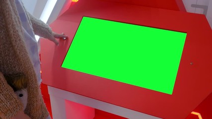 Woman looking at blank digital interactive green display kiosk at exhibition or museum with futuristic sci-fi interior. Mock up, copyspace, template, chroma key, green screen, technology concept