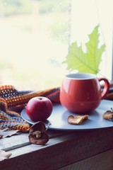 Bright cup with coffee on a window background, apples on a plate, warm scarf, leaves, autumn season, home comfort concept