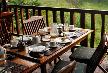 Wooden desk after morning breakfast - remains on plates and coffee cups, butts in ashtray, half empty milk jug, blurred green foliage in background - tropical holiday resort